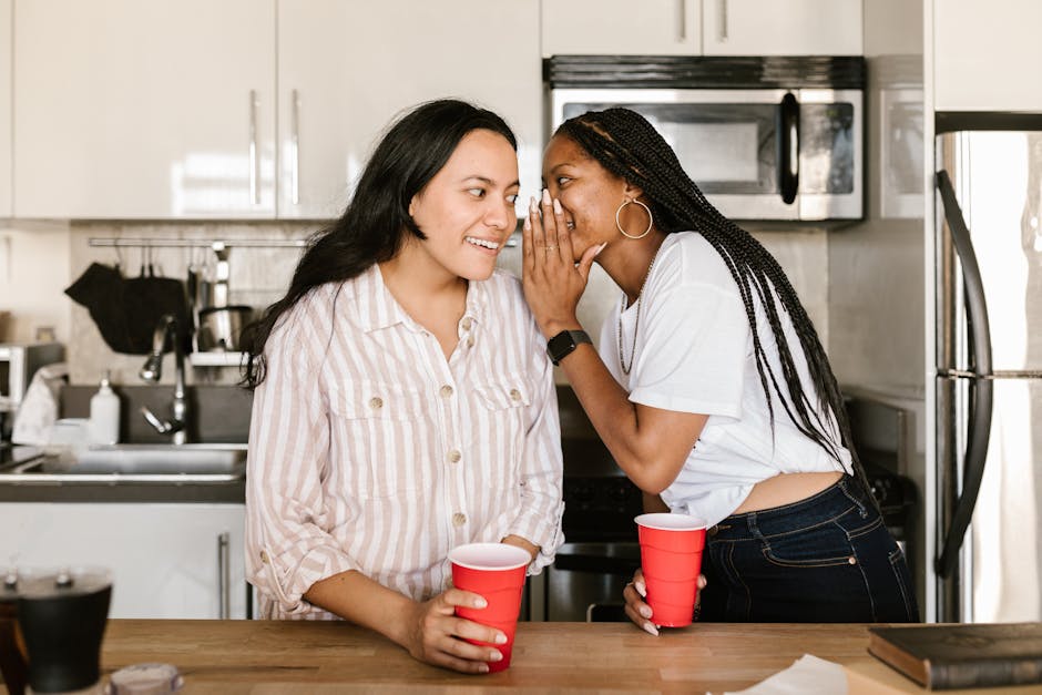 Two diverse women enjoying a candid moment and whispering secrets in a modern kitchen setting.