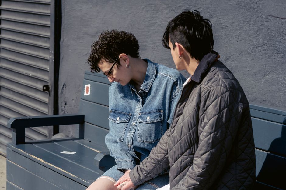 Side view of young androgynous woman with dark short hair holding hand of unhappy girlfriend sitting on bench on street