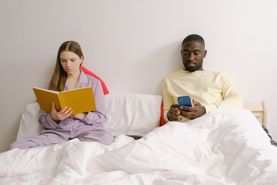 Concentrated woman with notebook sitting on bed near African American boy surfing cellphone while spending time together in light bedroom