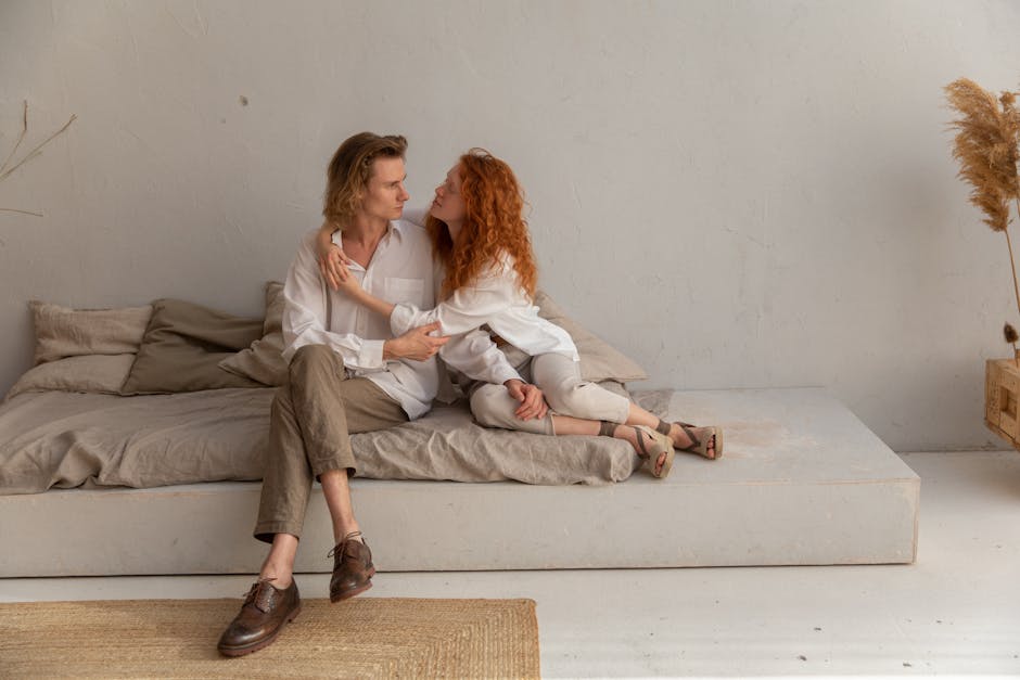 Full body of romantic couple hugging and looking at each other while sitting on comfortable mattress near wall in cozy room