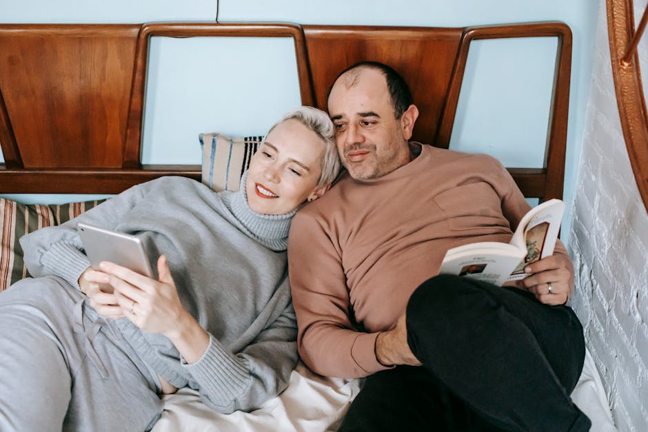 High angle of happy adult woman using tablet and cuddling with ethnic husband reading interesting book on bed at home
