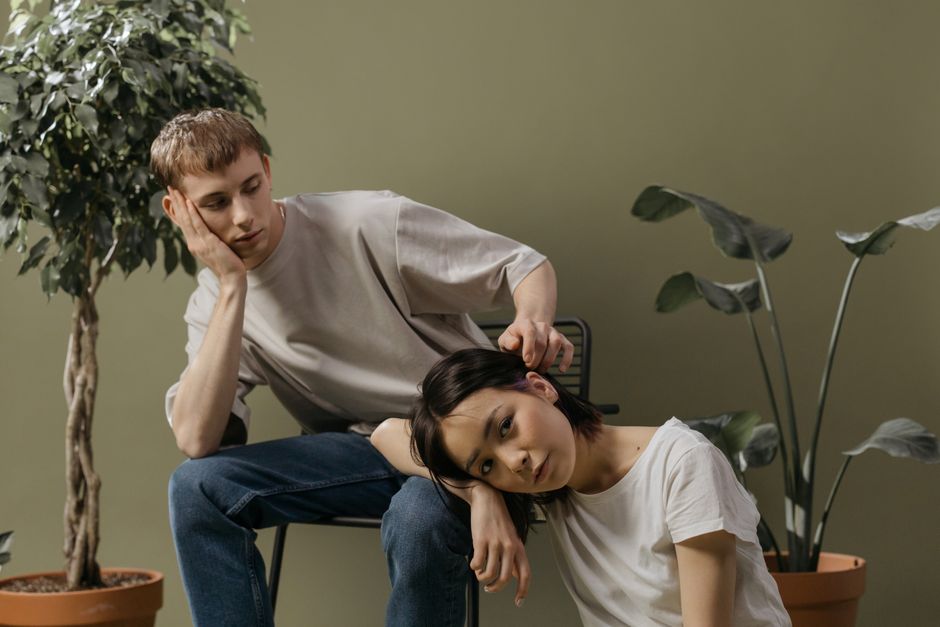 Two young adults in a minimalistic indoor setting surrounded by greenery, showing a moment of introspection.