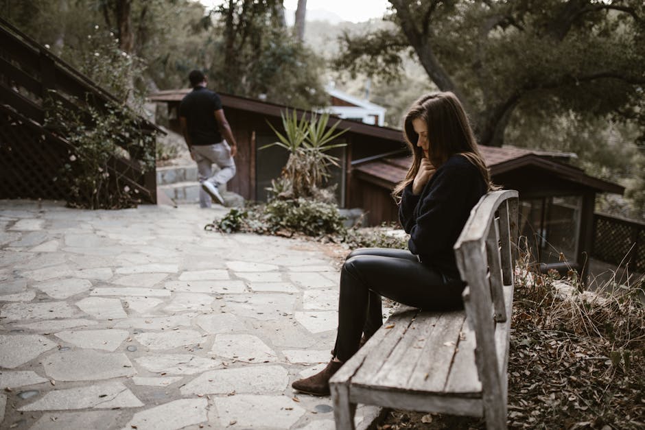 A woman sits pensively on a bench outside after a breakup, while a man walks away.