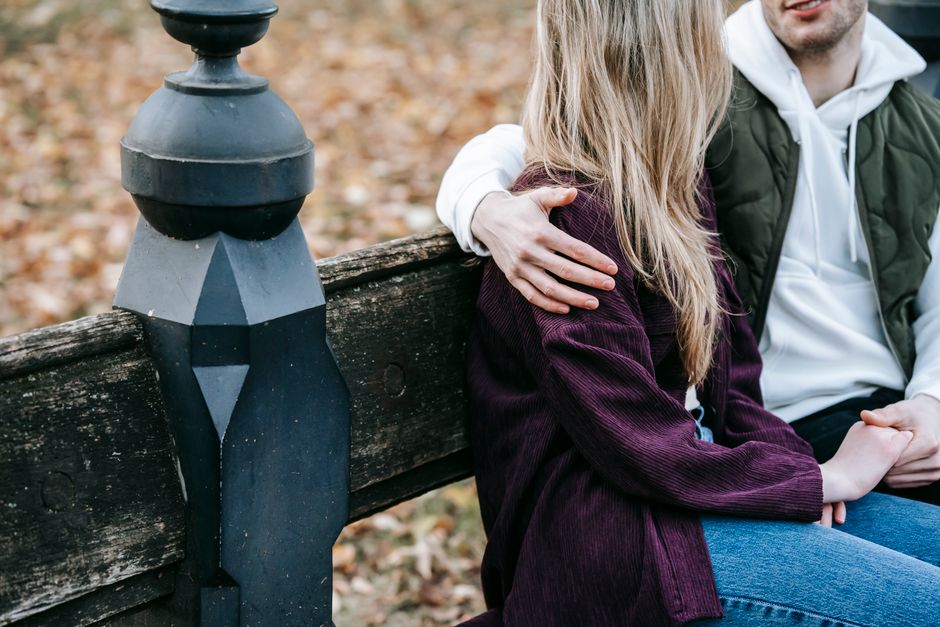 Crop anonymous loving couple resting on bench while holding hands in park with autumn leaves