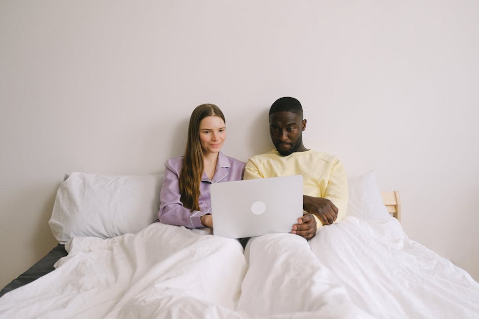 Interracial couple sitting in bed using a laptop, showcasing love and technology at home.